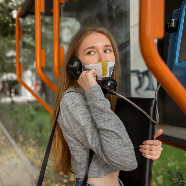 Mujer joven con máscara médica hablando por un teléfono público