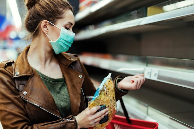 Mujer joven con máscara protectora y comprando comestibles en una tienda vacía en tiempos de epidemia de virus