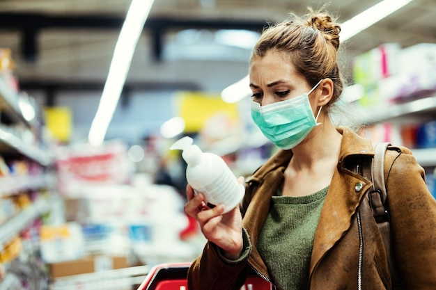 Mujer joven con máscara protectora leyendo la etiqueta en la botella mientras compra jabón de manos en el supermercado durante la epidemia de virus