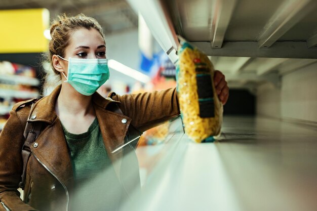 Mujer joven con máscara protectora mientras toma el último paquete de pasta en el supermercado durante la epidemia de virus