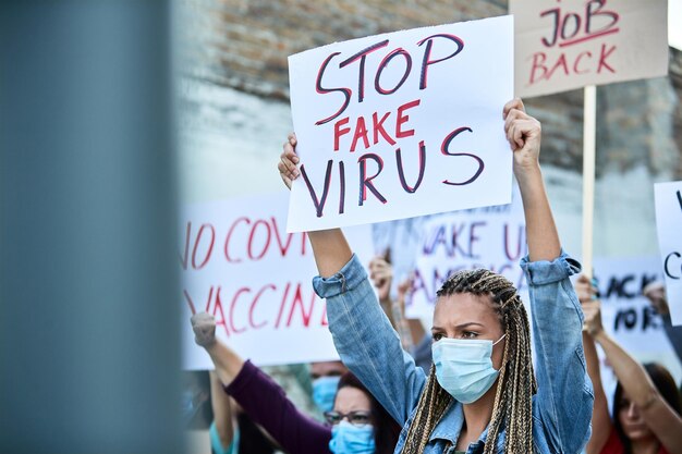 Mujer joven con mascarilla protectora que lleva una pancarta con la inscripción "detener el virus falso" mientras protesta con una multitud de personas contra los bloqueos debido a la epidemia de coronavirus