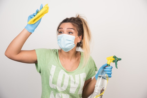 Mujer joven con mascarilla y suministros de limpieza en la pared blanca.