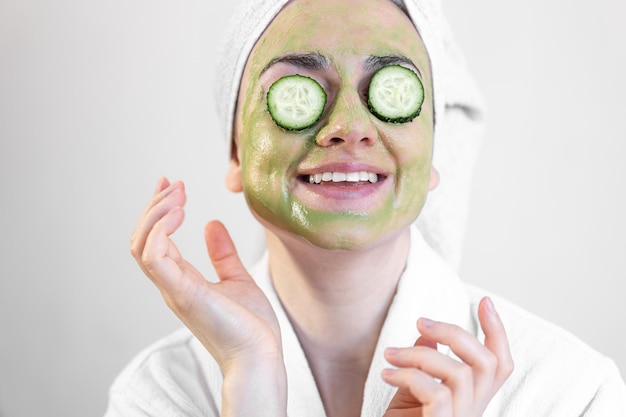 Mujer joven con mascarilla verde y pepinos frescos en una bata blanca