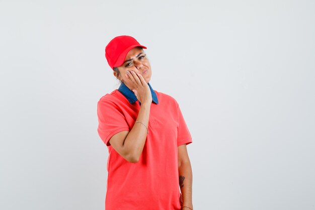 Mujer joven con la mejilla apoyada en la palma, con dolor de muelas en camisa roja y gorra y aspecto agotado. vista frontal.