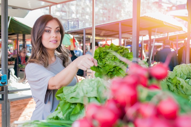 Mujer joven en el mercado. Feliz morena joven recoger algunas verduras. Retrato de mujer joven hermosa que elige verduras de hoja verde