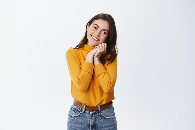 Mujer joven mirando algo adorable con afecto y ternura tomándose de la mano cerca de la mejilla y sonriendo mirando algo hermoso de pie contra el fondo blanco