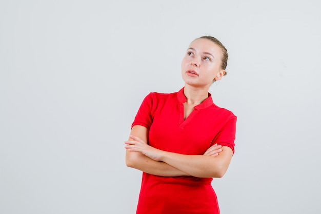Mujer joven mirando hacia arriba con los brazos cruzados en camiseta roja y mirando pensativo