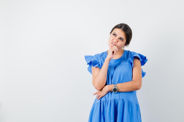 Mujer joven mirando hacia arriba con la mano en la barbilla en vestido azul y mirando pensativo