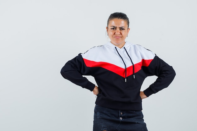 Mujer joven mirando a la cámara mientras sostiene las manos en la cintura en una colorida sudadera y parece descontento. vista frontal.