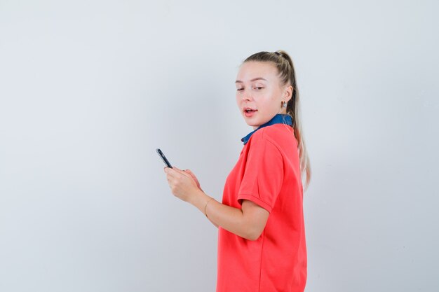 Mujer joven mirando a un lado mientras sostiene el teléfono móvil en camiseta y parece confiado.