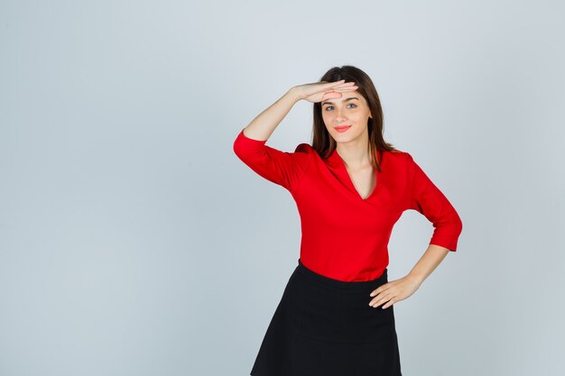 Mujer joven mirando lejos con la mano sobre la cabeza, sosteniendo la mano en la cadera en blusa roja