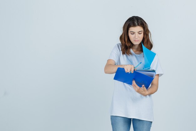 Mujer joven mirando notas en carpetas en camiseta blanca, jeans y mirando enfocado, vista frontal.