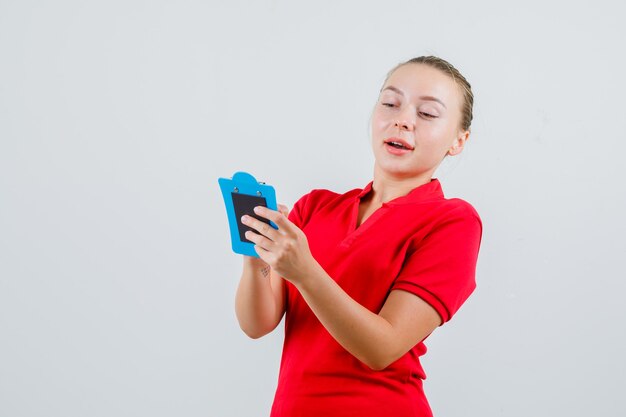 Mujer joven mirando notas en el portapapeles en camiseta roja y mirando positivo