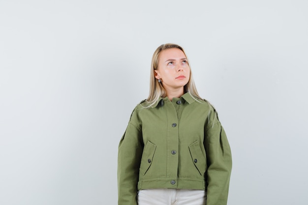 Mujer joven mirando a otro lado en chaqueta verde, jeans y mirando pensativo, vista frontal.
