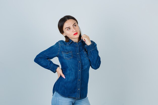 Mujer joven mirando a otro lado mientras mantiene la mano en la cadera en camisa de mezclilla y jeans y mirando pensativo