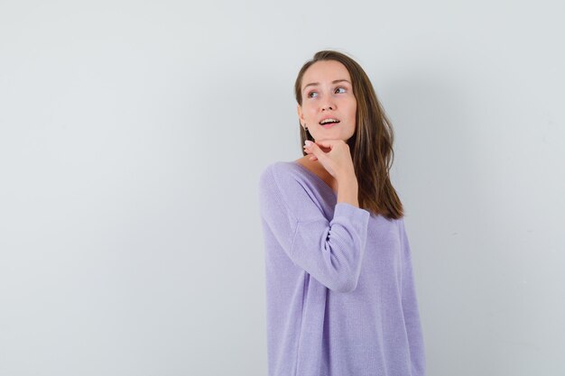 Mujer joven mirando a otro lado mientras sostiene la mano en la barbilla en una blusa lila y se ve impresionante. vista frontal. espacio para texto