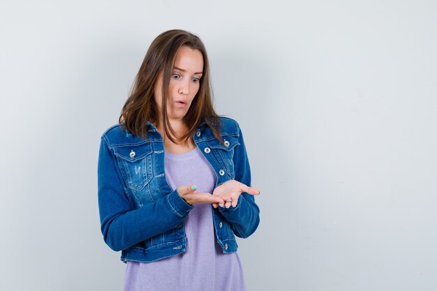 Foto gratuita mujer joven mirando sus manos abiertas en camiseta, chaqueta y mirando sorprendido, vista frontal.