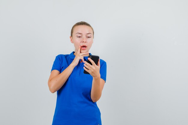 Mujer joven mirando teléfono móvil en camiseta azul y mirando asombrado