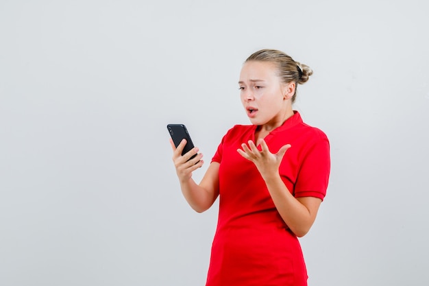 Mujer joven mirando el teléfono móvil en camiseta roja y mirando irritado