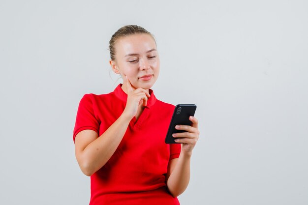 Mujer joven mirando el teléfono móvil en camiseta roja y mirando pensativo