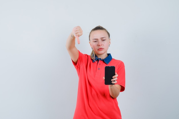 Mujer joven mirando el teléfono móvil con el pulgar hacia abajo en camiseta y mirando molesto