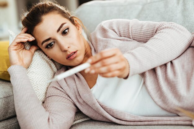 Mujer joven mirando el termómetro con incredulidad mientras mide la temperatura de su cuerpo en casa