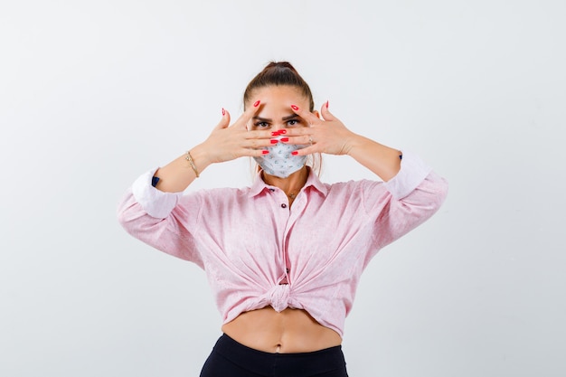 Mujer joven mirando a través de los dedos en camisa, pantalón, máscara médica y luciendo linda, vista frontal.