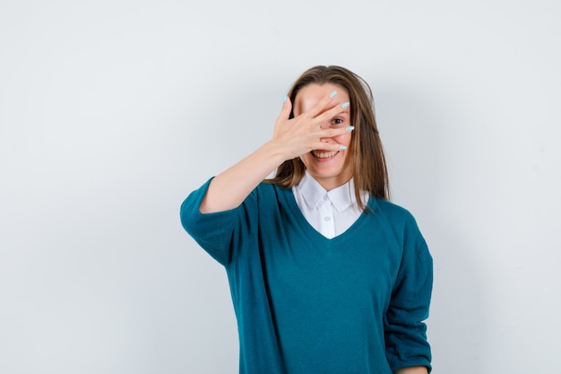 Mujer joven mirando a través de los dedos en suéter sobre camisa blanca y mirando alegre, vista frontal.