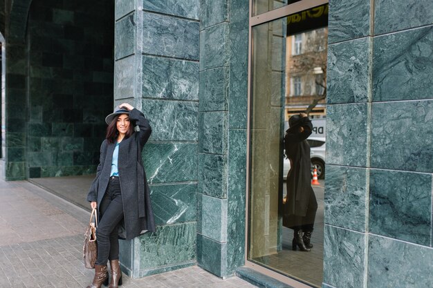 Mujer joven de moda en abrigo gris, sombrero caminando en la calle en el centro de la ciudad. Sonriendo, emociones verdaderas, estilo de vida con estilo, ropa de lujo, apariencia elegante.