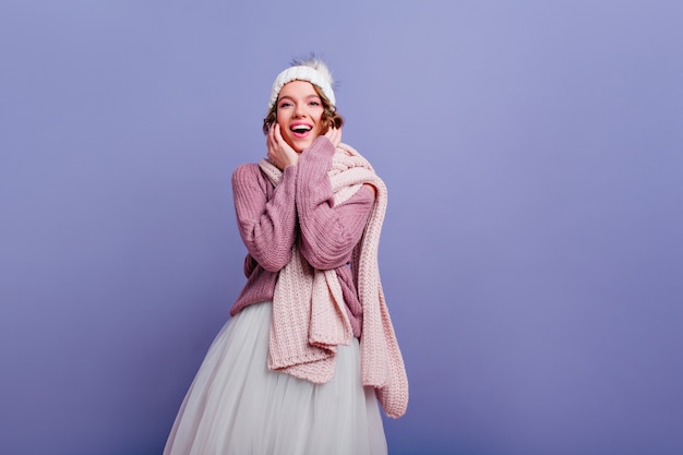 Mujer joven de moda en accesorios de invierno de moda posando Sonriente niña adorable en suéter de pie en la pared púrpura.