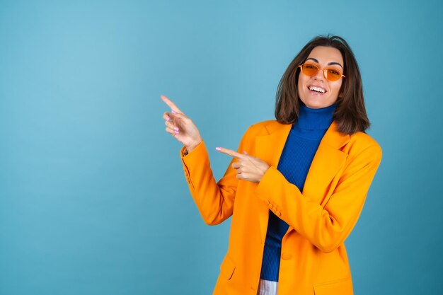 Mujer joven de moda en una chaqueta de gran tamaño y hasta la rodilla en una pared azul en elegantes gafas de color naranja brillante posando