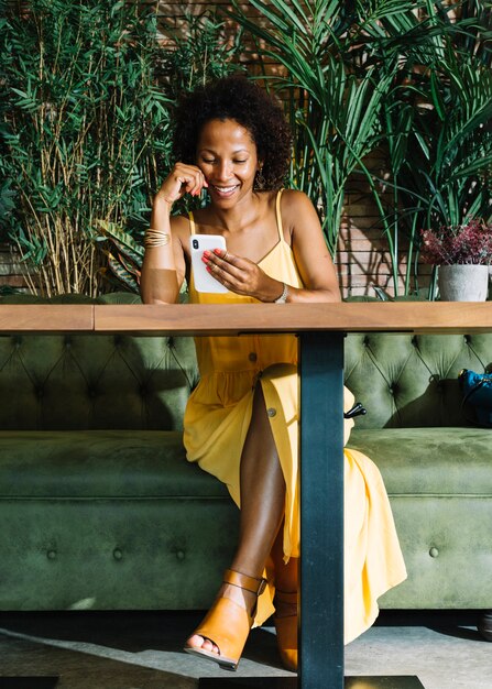 Mujer joven de moda feliz que se sienta en el restaurante que mira el teléfono elegante