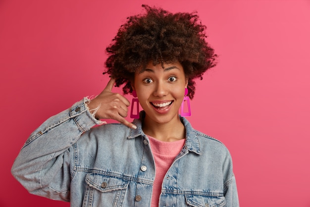 Mujer joven de moda hace gesto de teléfono, pide llamarla, expresa emociones positivas, viste ropa de mezclilla, sonríe feliz, aislado en la pared rosada. Llámame señal de vuelta. Lenguaje corporal