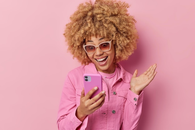 Mujer joven de moda positiva con cabello rizado enfocado en el teléfono inteligente recibe excelentes noticias en el mensaje navega en las redes sociales mantiene la palma levantada usa una chaqueta elegante aislada sobre fondo rosa