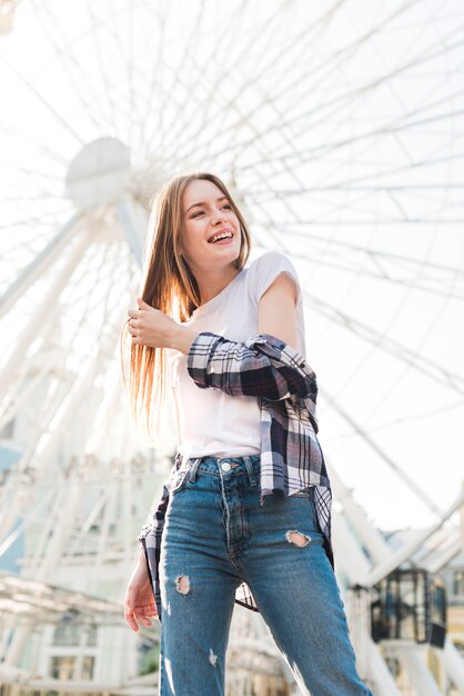 Mujer joven de moda que presenta delante de la rueda de ferris en el parque de atracciones