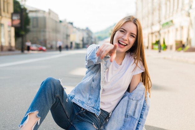 Mujer joven de moda que señala en la cámara que se sienta en el camino