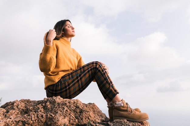 Mujer joven de moda que se sienta encima de la roca que disfruta de la naturaleza contra el cielo