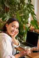 Foto gratuita mujer joven de moda sentada en un café y sonriendo a la cámara bebiendo café y usando una computadora portátil trabajando re