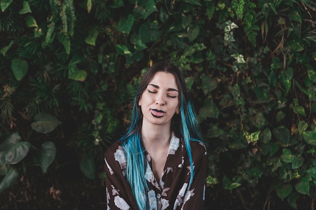 Mujer joven moderna con los ojos cerrados que muestran su lengua frente a una planta en crecimiento