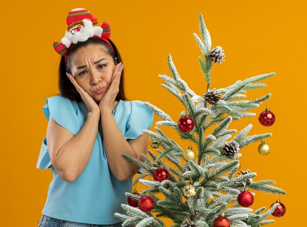 Mujer joven molesta en la parte superior azul vistiendo gracioso borde navideño en la cabeza mirando a la cámara con expresión triste de pie junto a un árbol de Navidad sobre fondo naranja