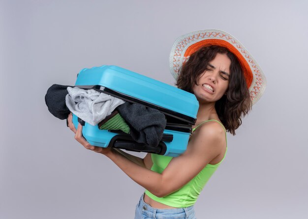 Foto gratuita mujer joven molesta que lleva sombrero y que sostiene la maleta llena de telas de pie en la vista de perfil en la pared blanca aislada