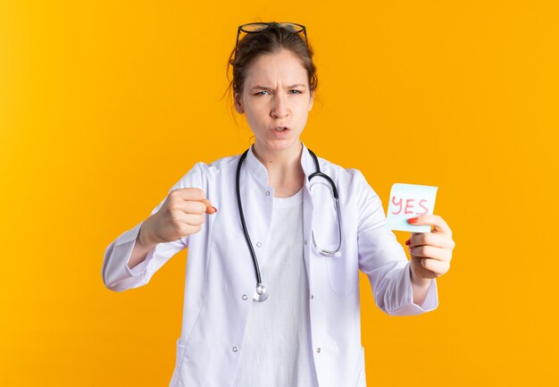 Mujer joven molesta en uniforme médico con estetoscopio sosteniendo una nota de sí y manteniendo el puño