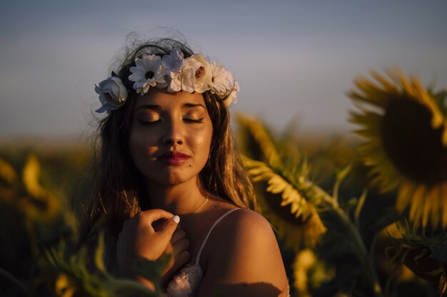 Mujer joven morena en una corona de flores con los ojos cerrados disfrutando del sol en un campo de girasol