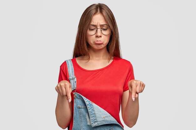 Mujer joven morena encantadora disgustada con expresión molesta, señala hacia abajo, nota algo desagradable, tiene aspecto molesto, usa un mono de moda con camiseta roja, posa contra la pared blanca