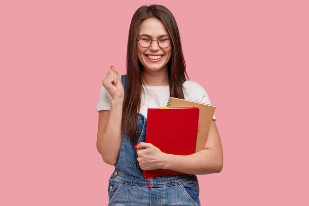 Mujer joven morena en overoles de mezclilla sosteniendo blocs de notas