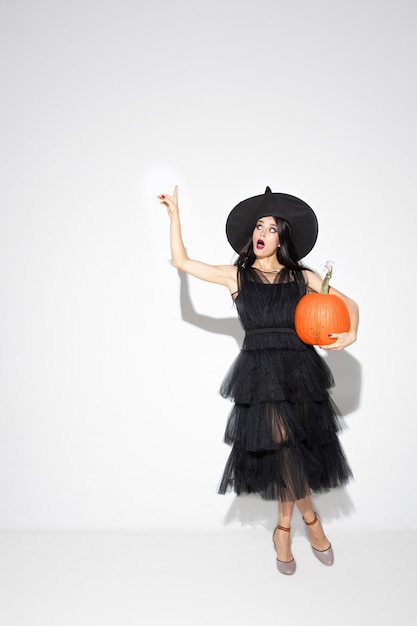 Mujer joven morena con sombrero negro y traje sobre fondo blanco. Modelo femenino caucásico atractivo. Halloween, viernes negro, cyber monday, ventas, concepto de otoño. Copyspace. Sostiene la calabaza, apuntando.