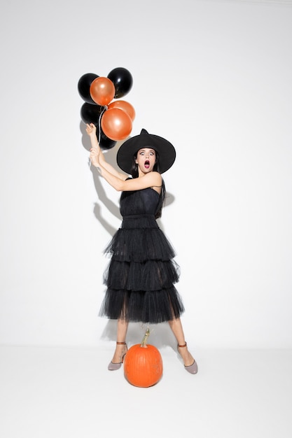 Mujer joven morena con sombrero negro y traje sobre fondo blanco. Modelo femenino caucásico atractivo. Halloween, viernes negro, cyber monday, ventas, concepto de otoño. Copyspace. Sostiene globos, sorprendido.