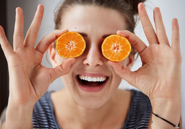 Mujer joven morena sosteniendo mandarinas en lugar de ojos, se ríe y disfruta de la vida