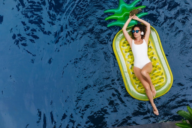 Mujer joven morena en traje de baño blanco escuchando música mientras nada en la piscina. Foto al aire libre de la encantadora dama morena divirtiéndose en el lugar de veraneo.