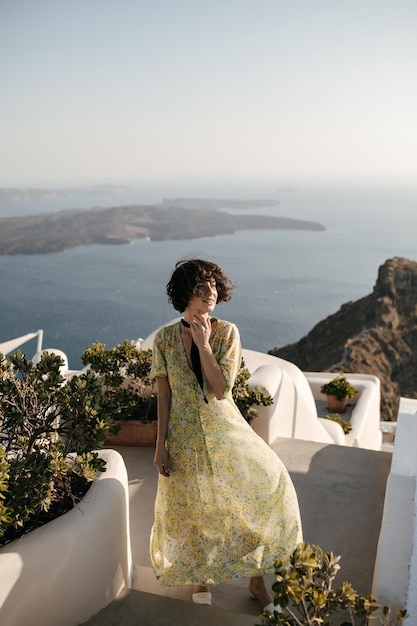 Mujer joven morena en vestido midi sonríe y disfruta de hermosas vistas al mar Dama de pelo corto rizado en traje floral posa en la terraza con vista al mar